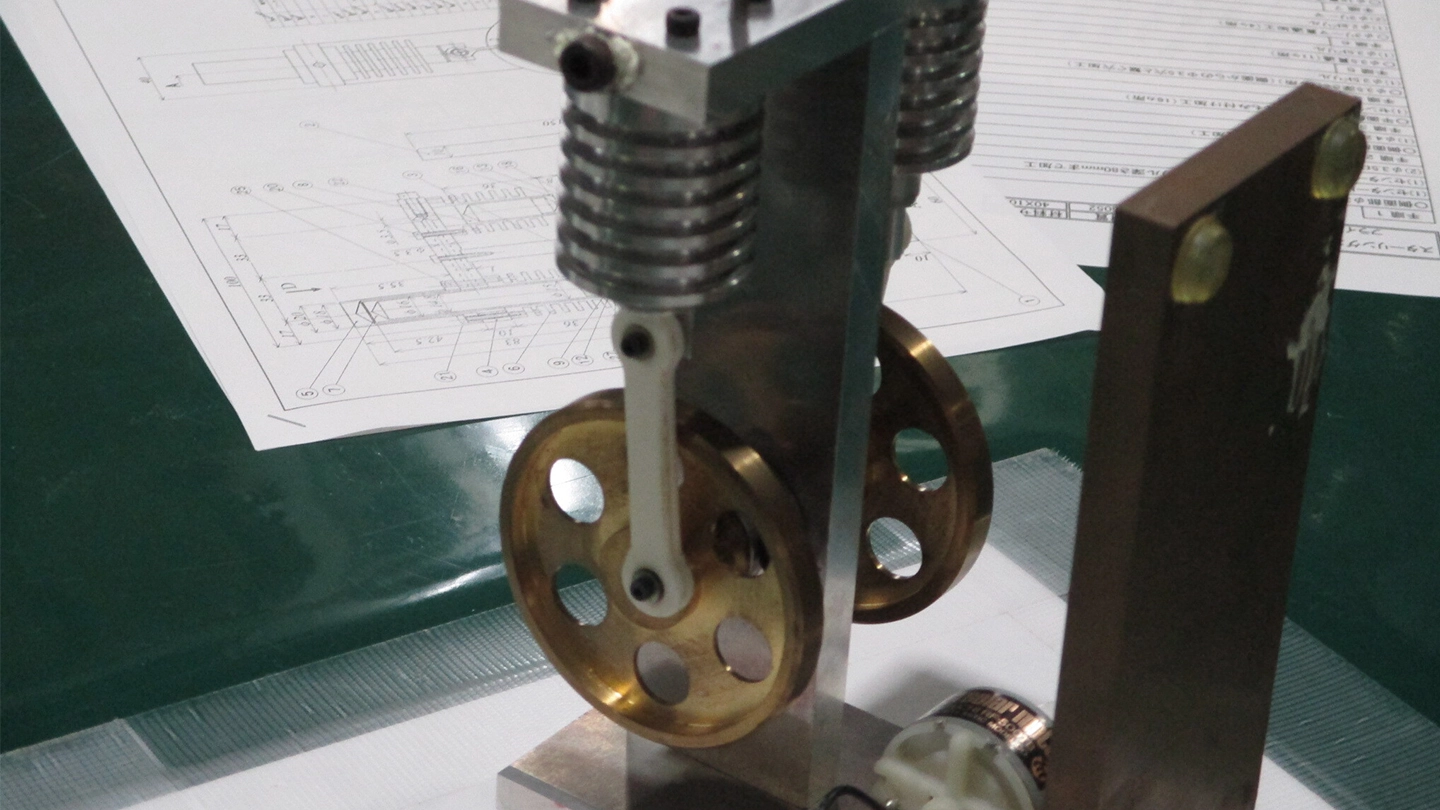 A small metal model of an engine. It features a cylinder, a crankshaft mechanism, and a brass-colored flywheel, with engineering drawings spread out in the background.